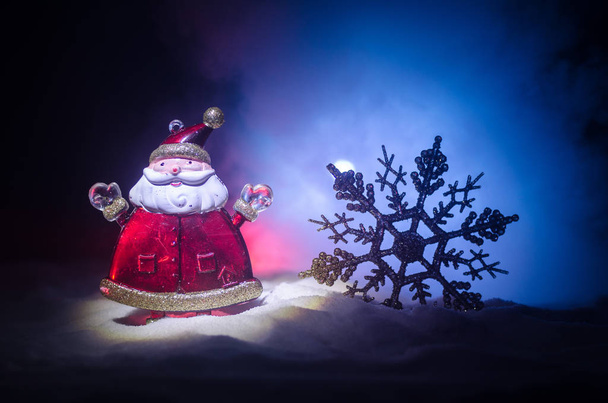 Happy Santa Claus Doll on Christmas time with tree and snow. Colorful bokeh background. Santa Clause and Merry Christmas model figure toy on dark toned foggy background. - Photo, Image