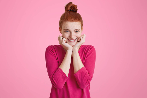 Headshot of pleased high spirit pretty female model with ginger hair knot, has warm pleasant smile, keeps hands under chin, wears pink sweater, isolated over blank copy space. Happiness concept - Foto, imagen