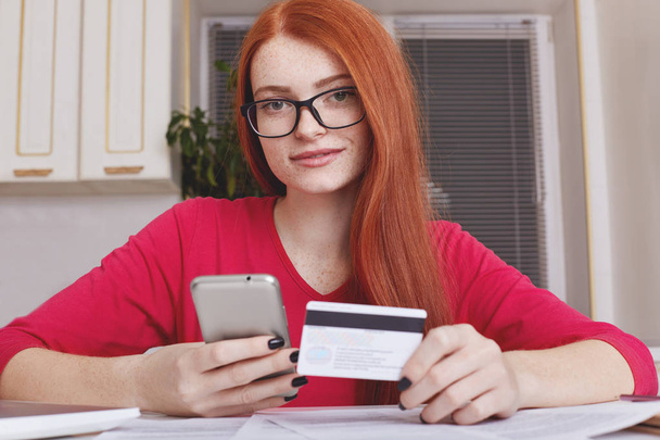 Beautiful redhaired female verifies credit card account balance on smart phone with mobile banking application, does shopping online. Mobile payments. Woman buys goods in internet enters security code - Fotó, kép