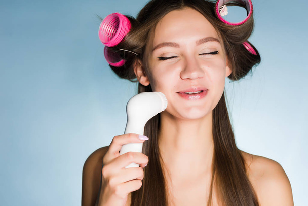 a satisfied young girl makes a deep cleansing of the skin on her face with an electric brush, on her head curlers - 写真・画像