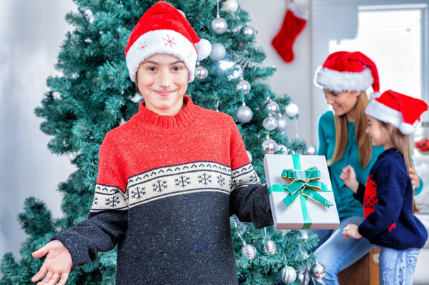 Νεαρό αγόρι κρατώντας το δώρο Χριστουγέννων με την οικογένειά του στο σπίτι. Happin - Φωτογραφία, εικόνα