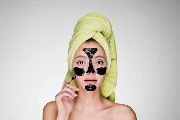 beautiful young girl with a green towel on her head applied a black cleansing mask to the problem areas on her face - 写真・画像