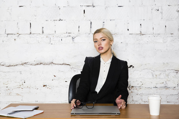 Young blond beauty businesswoman sitting at a office table with laptop, notebook and glasses in suit. Business concept. - 写真・画像