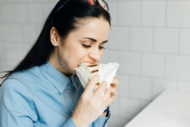 hungry young brunette girl in blue shirt eagerly eats sandwich in cafe - Fotó, kép