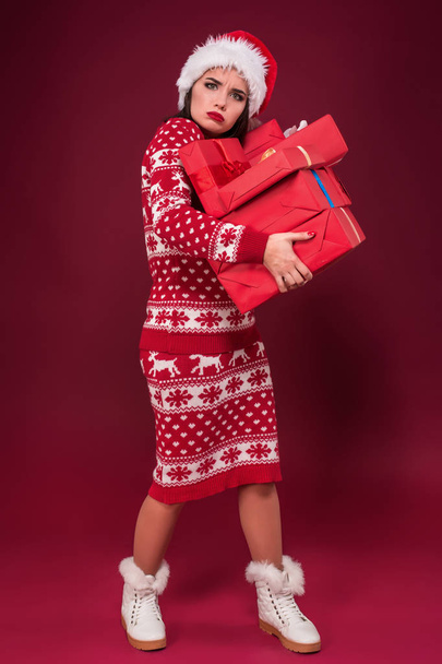 Νεαρή γυναίκα με το φόρεμα και Χριστουγεννιάτικο καπέλο κρατώντας κουτιά δώρων και εξετάζοντας τη φωτογραφική μηχανή σε κόκκινο φόντο - Φωτογραφία, εικόνα