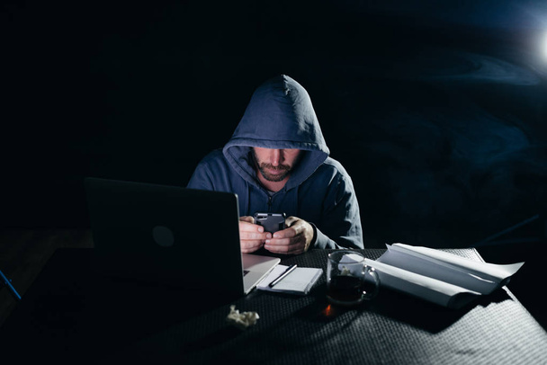 mysterieuze criminele man in een capuchon hacker, verbergt zijn gezicht, in de buurt van een laptop, schrijft iets op smarfone - Foto, afbeelding