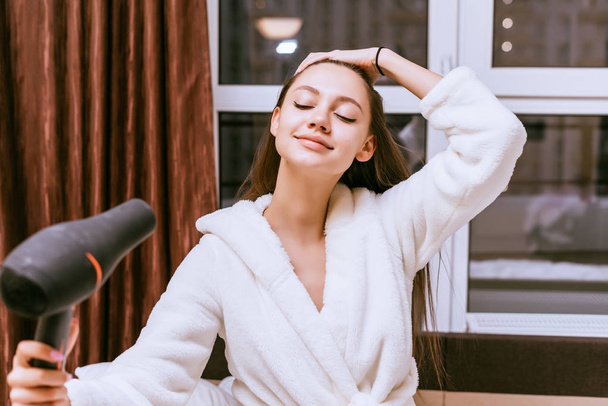 smiling girl in white bathrobe drying her long hair with a hairdryer - Foto, immagini