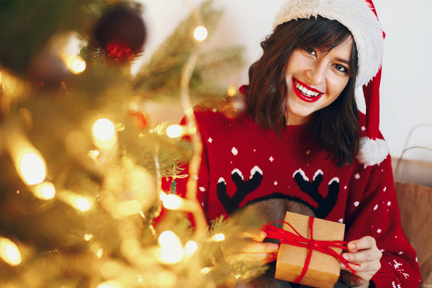 Žena v Santa hat drží stylový Dárkový box sedí pod vánoční stromeček s věnec světel - Fotografie, Obrázek