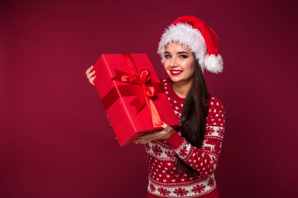 Jovem bela mulher segurando presente de Natal em suas mãos no fundo do estúdio vermelho
 - Foto, Imagem