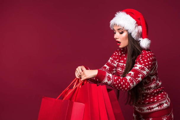 Jovem mulher bonita ou menina com cabelo longo bonito em chapéu de Santa e vestido de Ano Novo segurando pacote vermelho no fundo estúdio vermelho
 - Foto, Imagem