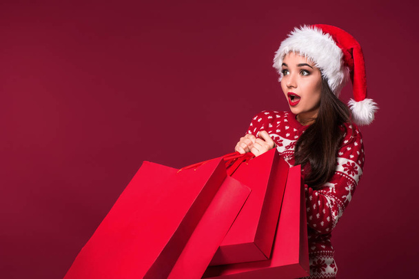 Young pretty woman or girl with long beautiful hair in Santa hat and New Years dress holding red package on red studio background - Φωτογραφία, εικόνα