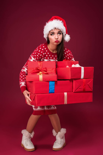 Young woman in dress and christmas hat holding gift boxes on red background - Fotografie, Obrázek