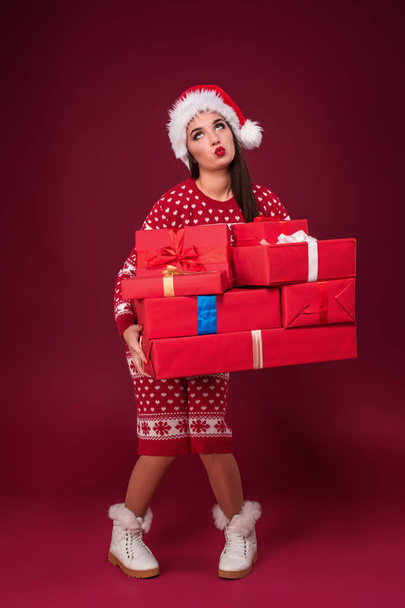 Young woman in dress and christmas hat holding gift boxes on red background - Fotó, kép