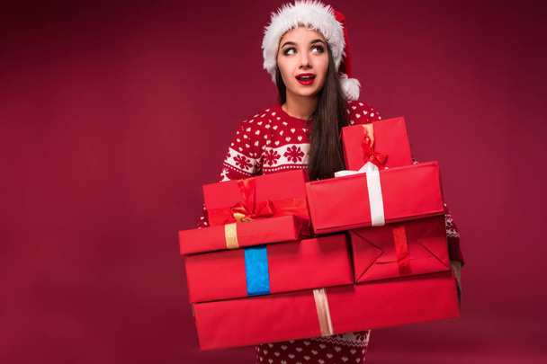 Young woman in sweater and christmas hat holding gift boxes on red background - Foto, Imagen