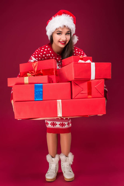 Bella donna stupita che tiene molti regali su sfondo rosso. Giovane donna in cappello di Babbo Natale guarda la fotocamera
 - Foto, immagini