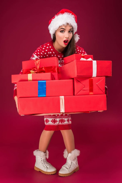 Amazed beautiful woman holding many presents on red background. Young woman in santa hat looks at camera - Foto, Imagem