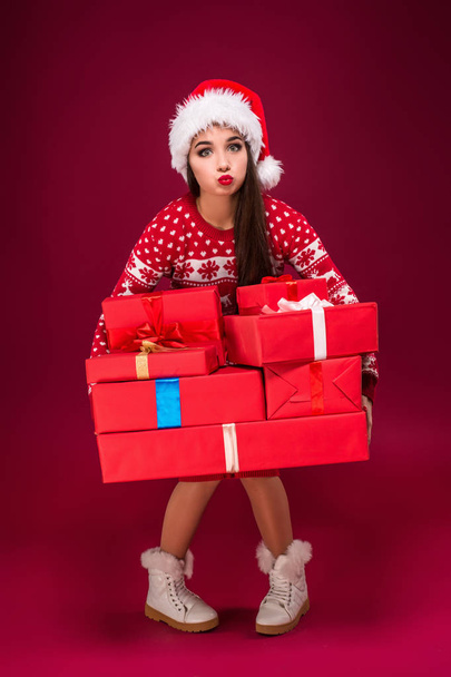 Amazed beautiful woman holding many presents on red background. Young woman in santa hat looks at camera - Photo, image