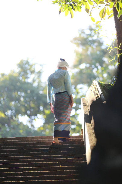 Porträt einer thailändischen jungen Frau in traditioneller thailändischer Kleidung, die im thailändischen Tempel mit Baum und Sonnenaufgang spaziert - Foto, Bild