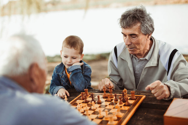 ενεργό συνταξιούχος senior ανδρών διασκεδάζοντας και παίζοντας σκάκι στο πάρκο, ξοδεύοντας το χρόνο με τον μικρό εγγονό - Φωτογραφία, εικόνα