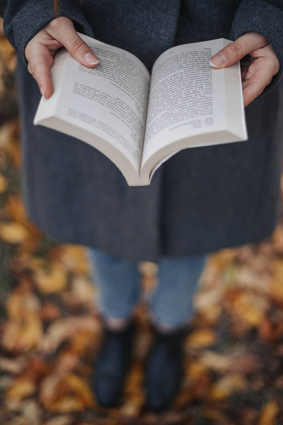 Female hands holding open book. Woman read book outdoor. Model wearing grey coat blue jeans and black boots.  - Zdjęcie, obraz