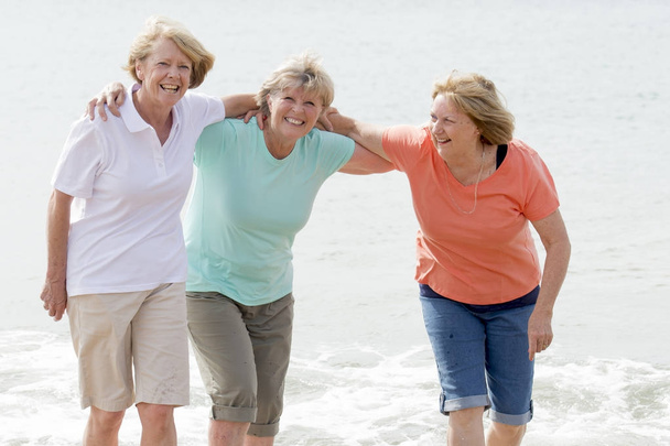 прекрасная группа из трех пожилых зрелых женщин на пенсии в возрасте 60 лет, веселящихся вместе счастливо гуляя по пляжу улыбаясь игриво
 - Фото, изображение