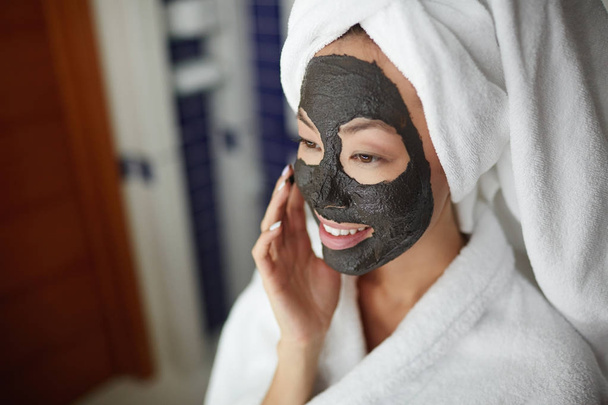 Portrait of beautiful Asian woman applying face mask during beauty treatment routine after shower, looking in mirror and smiling  - Photo, image