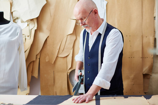 Portrait of old tailor cutting fabric on table while working in atelier shop - Fotoğraf, Görsel