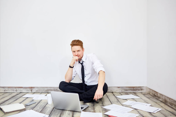 Nykyaikaisen nuoren liikemiehen muotokuva istuu ristissä lattialla toimistossa, työskentelee kannettavan tietokoneen kanssa ja katselee kameraa
 - Valokuva, kuva