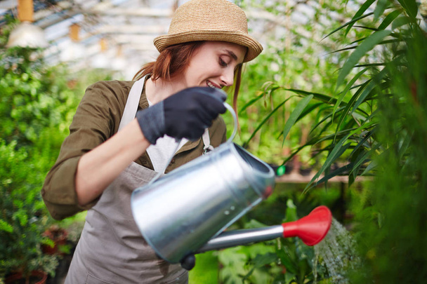 Retrato de vista lateral de una mujer joven con sombrero de paja disfrutando trabajando en el jardín del árbol, regando las plantas con latas de metal y sonriendo
 - Foto, Imagen