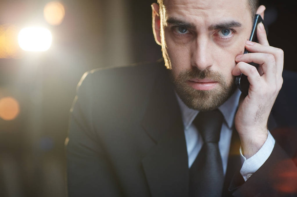 Retrato de hombre de negocios moderno con traje formal negro mirando a la cámara mientras habla por teléfono móvil contra fondo negro con destello de lente
 - Foto, imagen