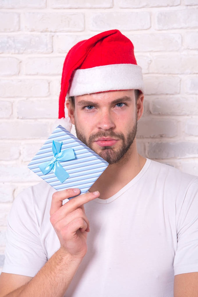 Macho in santa hat with present on white brick wall. Boxing day concept. Man hold xmas box with blue ribbon. Christmas, new year preparation and celebration. Gift giving, holiday greeting. - Foto, imagen