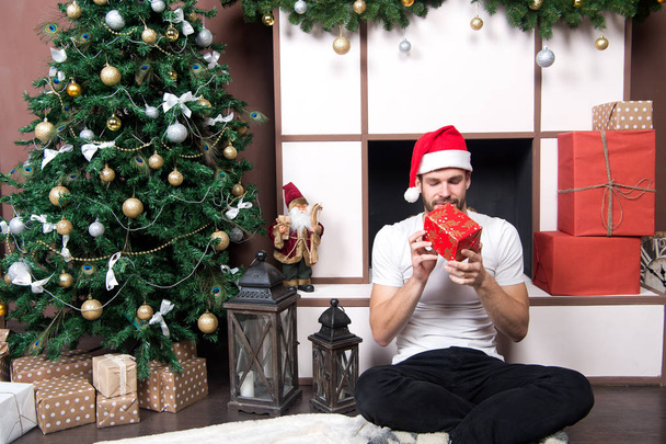 Macho with closed eyes hold present box at Christmas tree. Man in santa hat sit at fireplace. Boxing day concept. xmas and new year room interior. Winter holidays preparation and celebration. - Photo, image