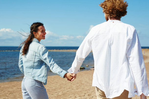 Visão traseira do jovem casal feliz durante o passeio na praia de areia no dia de verão
 - Foto, Imagem