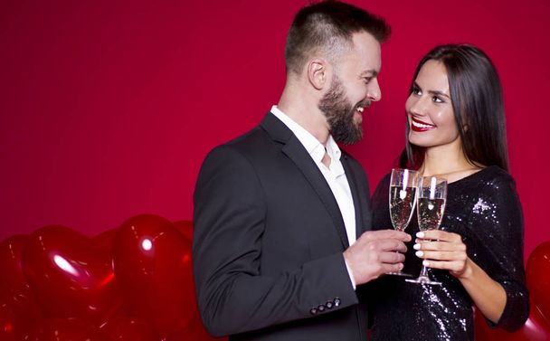 Lovers rest. Gentle stylish beautiful smiling couple of lovers with glasses of champagne on a red background with balls in the shape of heart. Women's Day or March 8th.  - Fotó, kép