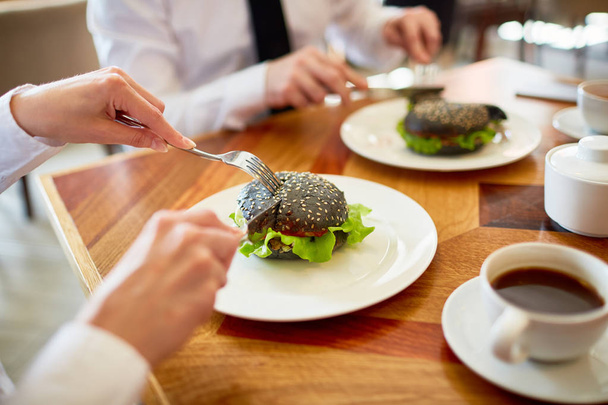 Primer plano de compañeros irreconocibles en ropa formal sentados en la mesa de la cafetería y disfrutando de hamburguesas apetitosas y té de hierbas
 - Foto, imagen