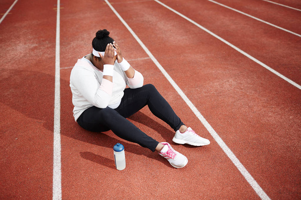 High angle view of frustrated overweight woman in sportswear sitting on running track with her head in hands, feeling exhausted - Zdjęcie, obraz