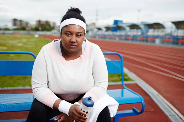 Chica de gran tamaño cansado en ropa deportiva sentado en el banco con toalla y botella de agua
 - Foto, Imagen