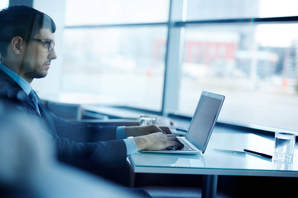 Profil Zobacz koncentratu młody biznesmen w okularach, siedząc przy biurku i pisanie list z odpowiedzią do jego partnera na nowoczesny laptop, zobacz przez ściany szklane - Zdjęcie, obraz