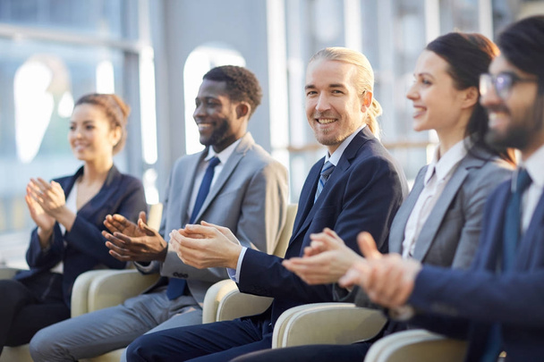 Grupo multi-étnico de empresários sorridentes sentados em fila no salão de vidro moderno e palmas, foco em jovens empresários alegres no centro
 - Foto, Imagem
