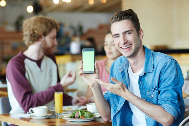 Красивый молодой человек с широкой улыбкой позирует для фотографии, указывая на пустой экран современного смартфона, его друзья живо общаются друг с другом и наслаждаются обедом в уютном кафе
 - Фото, изображение