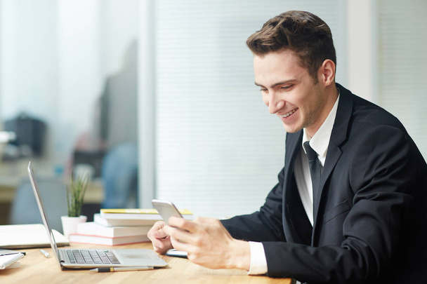 Profiel te bekijken van jonge witte kraag werknemer texting met zijn collega op smartphone glimlachen terwijl u zit in de voorkant van de laptop bij moderne kantoor, taille-staand - Foto, afbeelding