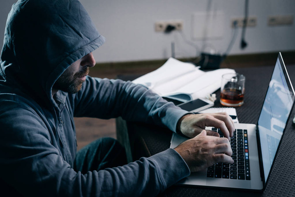 mysterious criminal man in a hood hacking something on a laptop - Foto, Imagen