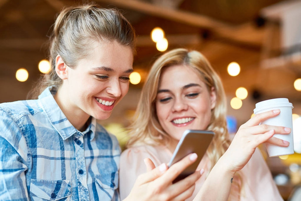 Vrolijke jonge vrienden met een charmante glimlach kijken naar grappige video op smartphone en genieten van cappuccino tijdens de vergadering op gezellige koffiehuis - Foto, afbeelding