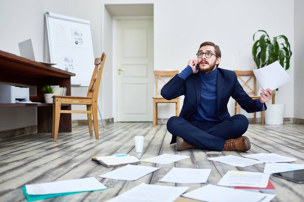 Portrait of bearded man speaking by phone looking troubled and sorting documents sitting on floor in office - Foto, afbeelding