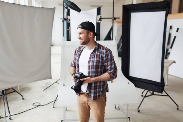 Mosolygó szakállas fotós viselő portréja kockás ing és a baseball sapka keres el, miközben rövid szünet a munkából, belső tágas photostudio háttér - Fotó, kép