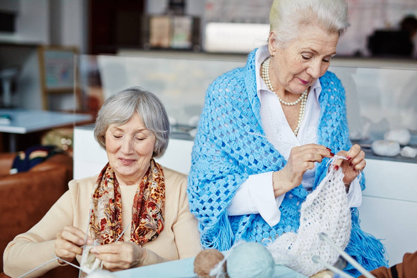Hermosa mujer mayor en azul chal de punto ganchillo mientras que su mejor amigo alegre sentado a su lado y tejiendo con ayuda de cuatro agujas
 - Foto, imagen