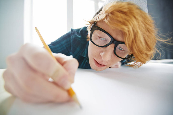 ビーニー帽子とデスクに近い傾いたして焦点を当て鉛筆でメガネを身に着けている若い創造的な赤髪の男の肖像 - 写真・画像