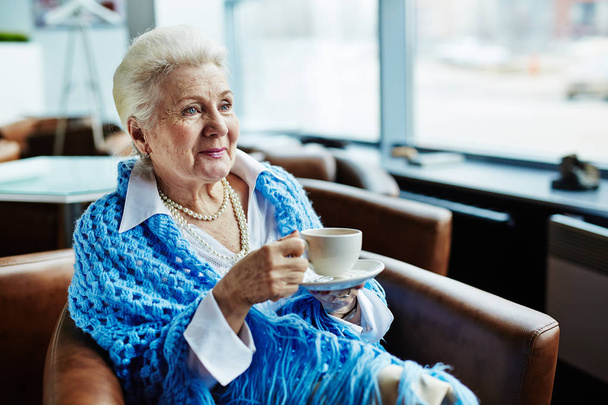 Portrait of elegant-looking elderly woman with cornflower blue knitted shawl on shoulders holding cup of delicious coffee in hands, sitting in comfortable armchair and enjoying picturesque view from window  - Zdjęcie, obraz