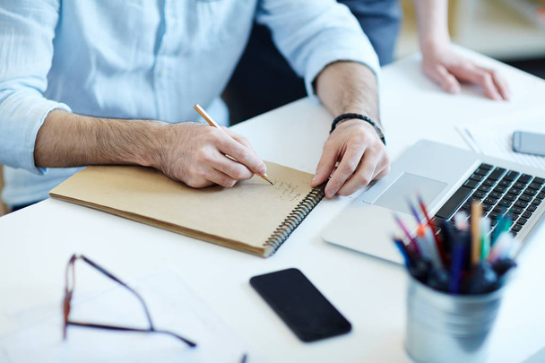 Крупный план мужских рук, пишущих в блокноте во время работы за столом в офисе
 - Фото, изображение