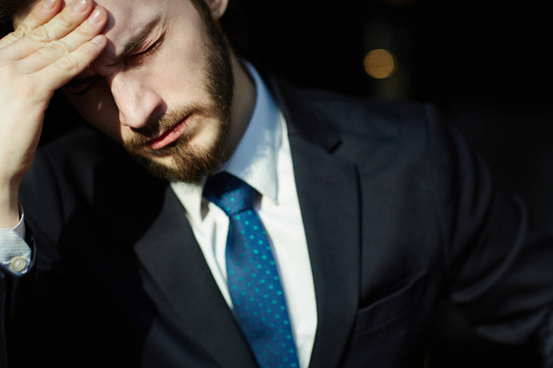 Sunlit head and shoulders portrait of handsome bearded man wearing elegant business suit, closing his eyes and rubbing forehead trying to relieve tension and headache - Foto, afbeelding